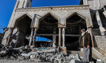 A bearded man in a cap stands before the heavily damaged Al-Huda Mosque against the backdrop of a blue sky. Within the rubble of the mosque, children have lined up to look at the damage left behind