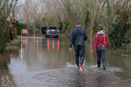 A young man and woman, seen from behind, walk through water on a flooded road; he wears full-length waders and she has black wellies. A pickup truck is parked the other side of the water in the tree-lined lane and a sign indicates the road closure.