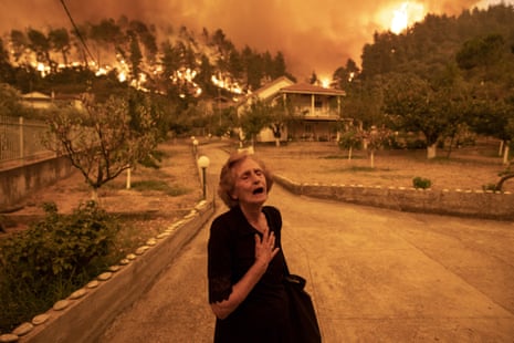 An older lady clasps her chest and shouts while fires rage in the background behind a large house in Greece
