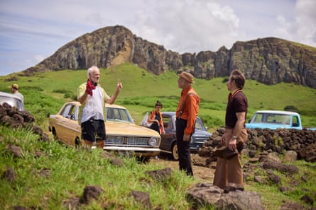 Martin McDonagh gestures with his arms to John Malkovich and Sam Rockwell next to vintage cars with mountains in the background