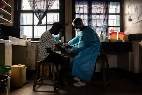 A healthcare worker in a blue gown and mask draws blood from a patient's arm in a clinical setting
