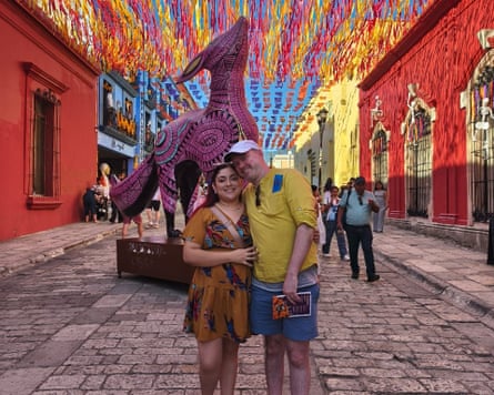 Man in a yellow shirt and blue shorts embracing a woman in an orange patterned dress on a street covered in purple and yellow hanging decorations