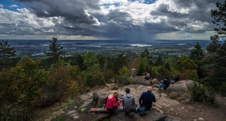 People picnic on a hill in the foreground with trees then in the distance there is a view of a sprawling city, stretching to a fjord