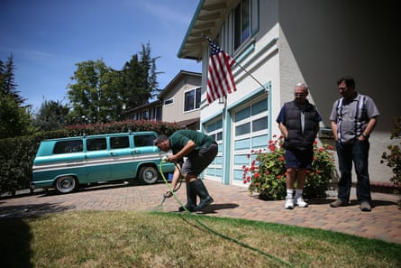 Two men watch as a third paints arid grass dark green with a spray gun outside a house in California