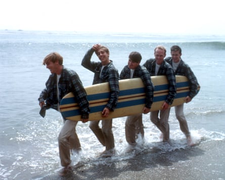 They walk along the beach holding a surfboard