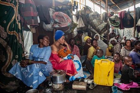 Women and small children sit surrounded by jerrycans, cooking pots and bedding in a shed