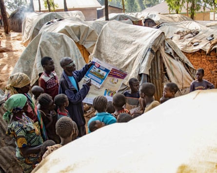 A man holds up an information sheet while speaking to a small crowd of people surrounded by tents.