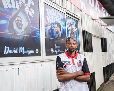 Benson Ngari poses for a photo beside posters on a wall showing top darts players.