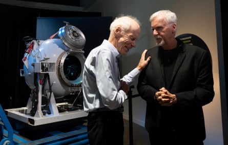 Two elderly white-haired men chat and laugh together next to a submersible pod in a museum