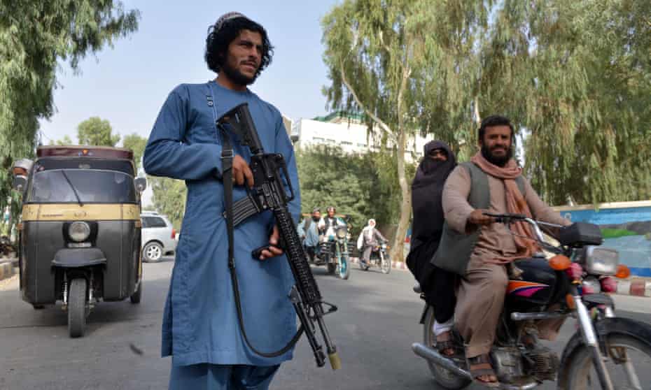 A young bearded man in a blue outfit with an assault rifle stands in the middle of traffic