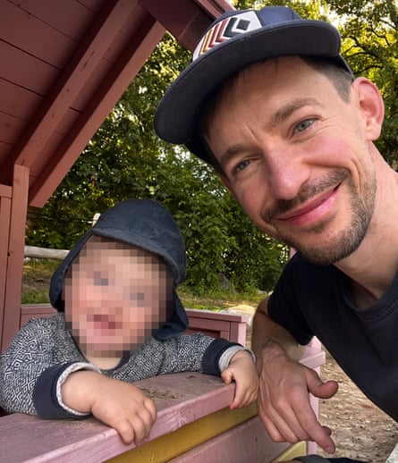 Simon Montgomery smiles beside his toddler, wearing a sun hat, at a playground
