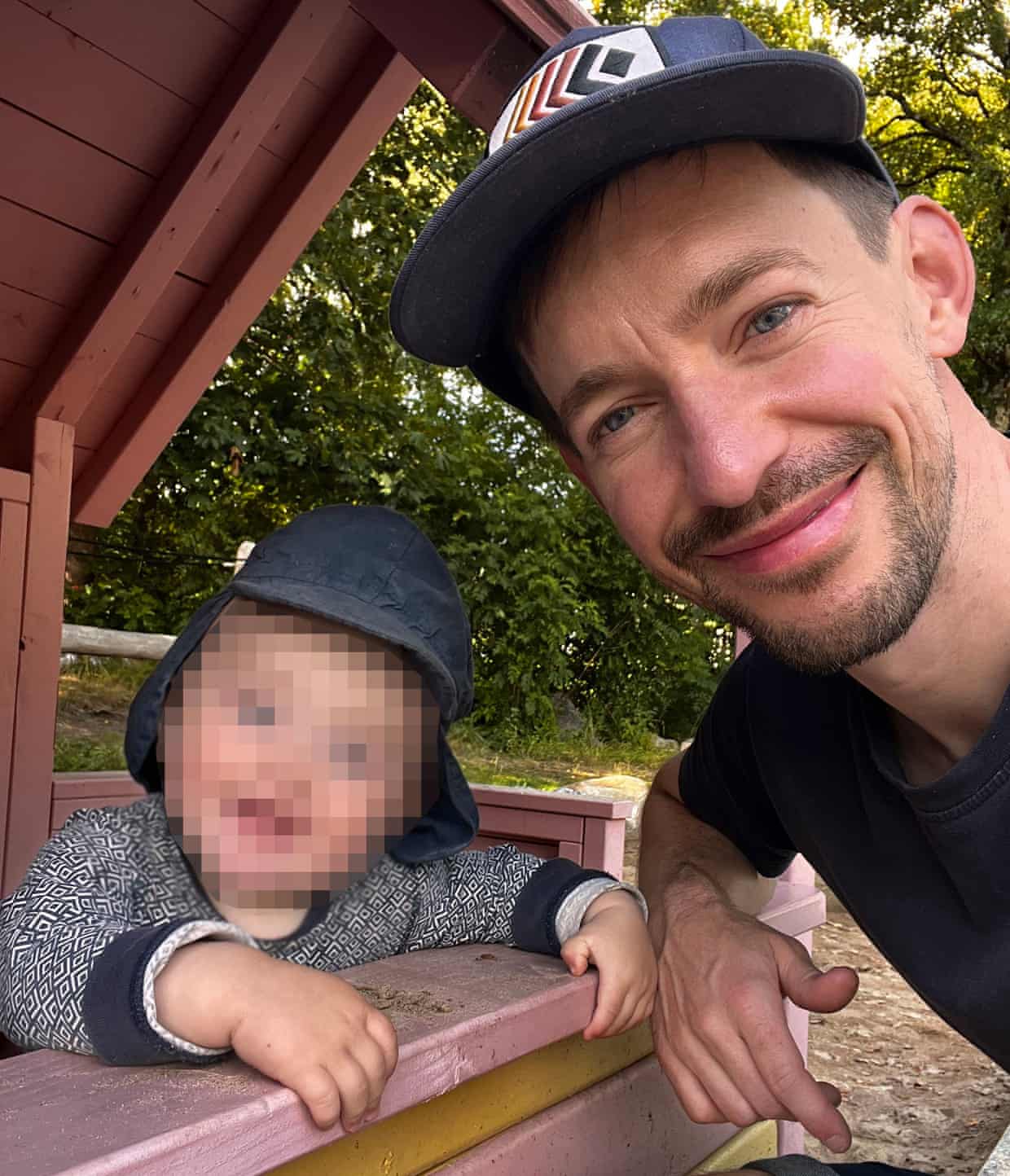 Simon Montgomery smiles beside his toddler, wearing a sun hat, at a playground