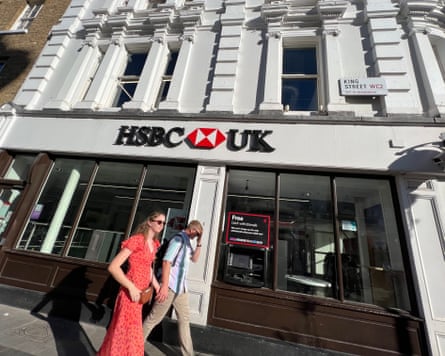 Two people, one in a red polka dot summer dress, cast a shadow on the side of a branch of HSBC in London on a sunny August day