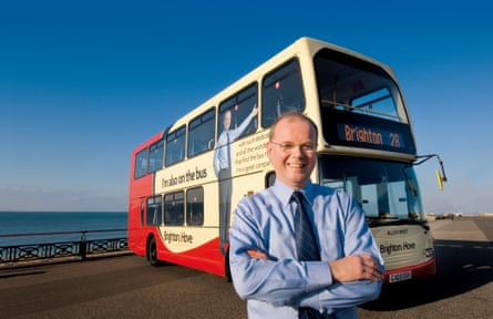 a man in a blue shirt with his arms crossed standing in front of a red and cream double decker with a bright blue sky behind him