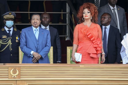 An old man and a woman in a huge red wig stand with military officers and officials on a podium