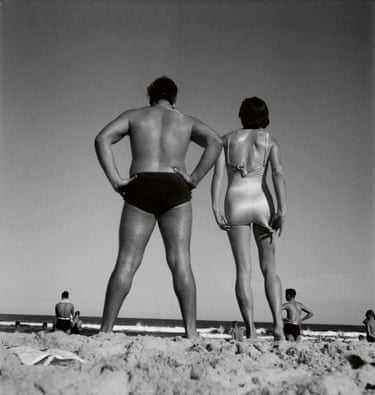 Black and white photograph of a man and a woman on a beach, wearing bathers, seen from behind