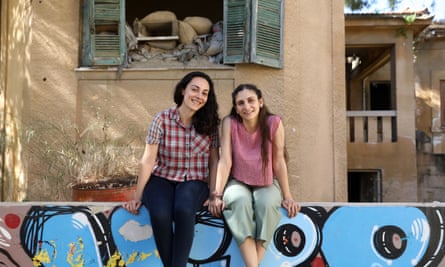 Feray Yalcuk and Maria Loucaidou sitting side by side and smiling, on a wall with grafitti on it and with a shabby building with broken shutters behind them