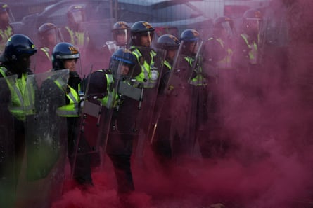 Riot police stand guard in a cloud of red smoke from a flare