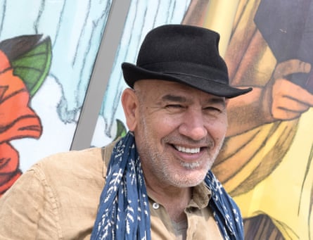 Rashid Masharawi, in a fedora hat with a long scarf, grins in front of a mural