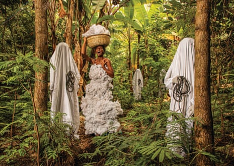 A woman in a white dress of cotton bolls stands with a basket on her head in a forest appearing to scream with three figures draped in white sheets and holding ropes stand nearby
