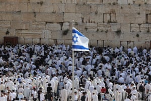 Hundreds of people in white shawls facing the Western Wall, as an Israel flag flies on a pole