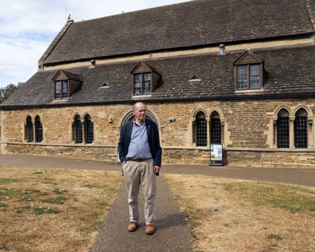 An older man stands in front of a pretty building in a village.