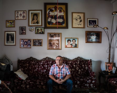 A man sits on a sofa indoors with a wall of pictures behind him