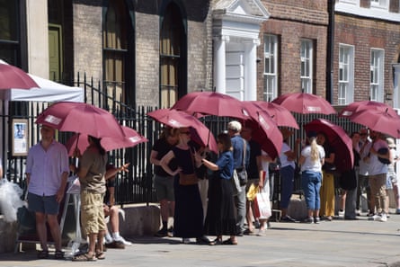 A line of people outside the museum, a historic brick building with pillars, stand under large dark red umbrellas.