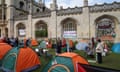 Students stand among tents pitched on a lawn outside a gothic-style university building