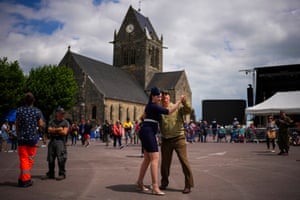 A couple in second world war costume dance in Sainte-Mère-Église town square with a church behind them