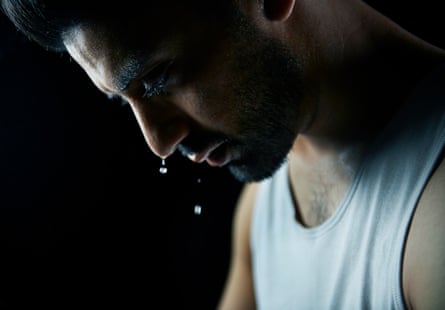 A close up profile of a man looking downward in a dark room in a white vest – with drops visibly falling from his depressed looking face