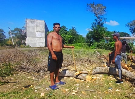 Two topless men, one holding gan axe, stand next to a small felled tree in a sunny field