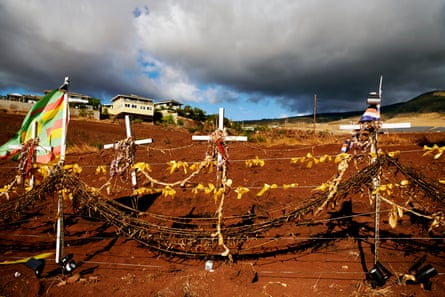 Thin white crosses with what appear to be yellow ribbons and small colorful flags flapping in wind, under storm clouds with some blue in the sky, on brown ground.