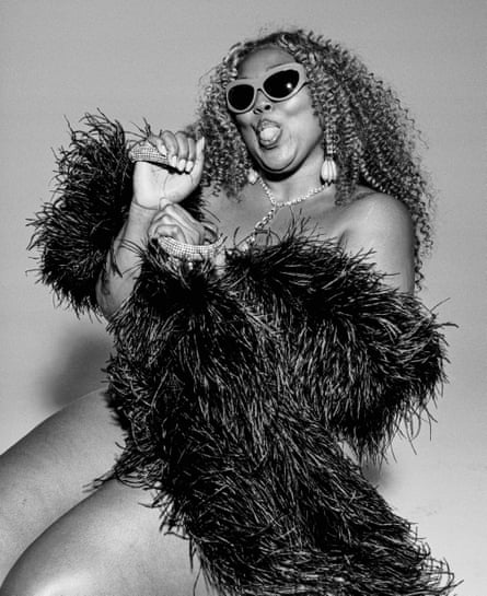 A black and white shot of Lizzo with only a big black feather bower around her, wearing shades and smiling while sticking her tongue out at the camera