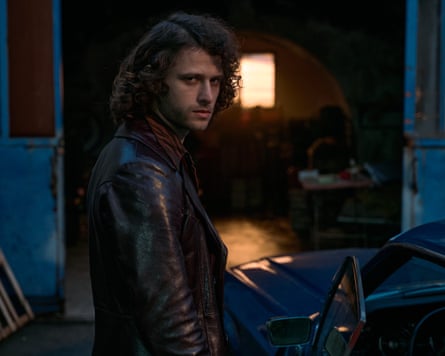 A long haired man in a leather jacket gives a sideways glance as he stands by the front of a sports car in the half light