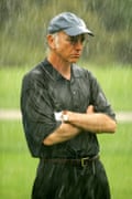 a man wearing hat and glasses, standing on a golf course while it rains