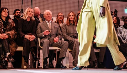Laura Weir, King Charles and Stella McCartney sit and watch a model walk past in a yellow suit and coat
