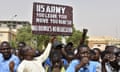 A group of Nigerien men with very short hair, wearing powder blue and white linen shirts, hold a brown and white handpainted sign that says "US Army, You Leave, You Vanish, No Bonus, No Negotiation."