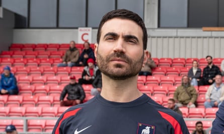 Man with beard wearing blue and red shirt in front of red bleachers.