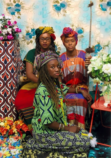 Three women in colourful headwraps and patterned clothing pose together amid flowers and fabric