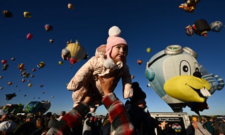 A cute infant in pink cold-weather gear is in the center of the frame, looking out over heads, with the sky behind her dotted with hot-air balloons, including one that looks like a cartoon chipmunk with aviator sunglasses.