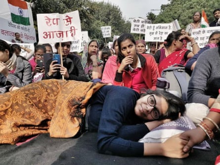Swati Maliwal lying on the ground under a blanket and with pillow surrounded be seated women holding placards, with one woman holding her hand.