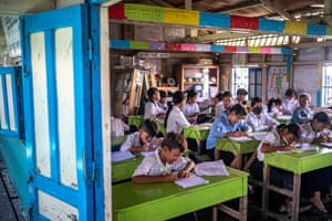 Children in school uniform sit with paper and writing instruments at green desks in a classroom