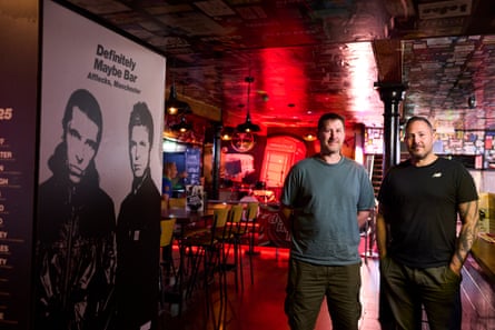 Two men in T-shirts standing in a bar next to a massive poster of Liam and Noel Gallagher