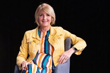 Kaye Adams sits in a chair wearing a yellow jacket and striped dress
