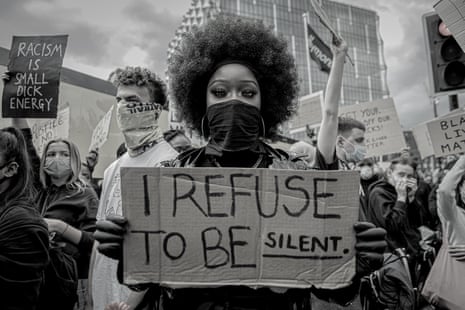 A young black protester with a big 'afro' hairstyle and a black bandana over her face holds a cardboard sign to the camera reading "I refuse to be silent"