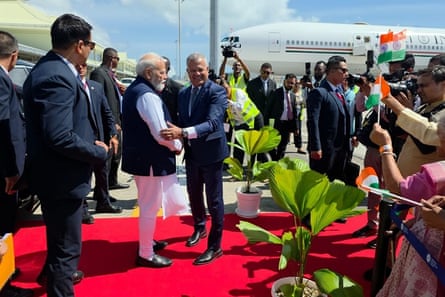 Modi stands on a red carpet at the airport with a plane in the background as officials stand by and people wave Indian flags.
