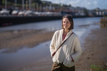 Emma Roberts stands by the side of the river with boats moored on the other side in the background. It is low tide and there are mudbanks.