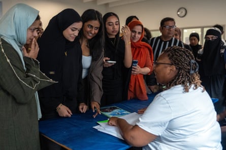 A group of girls, most wearing headscarves, stand around a table as a woman hands out their results.