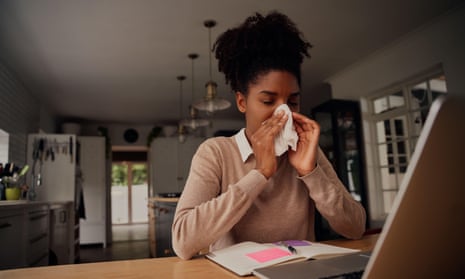 Young woman suffering from the cold virus works at a desk and laptop while blowing her nose